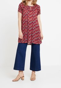 Rød tunika med et geometrisk mønster, løs silhuet, korte ærmer, parret med brede mørkeblå jeans og beige sandaler med remme.