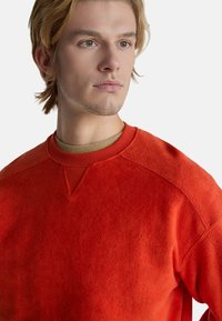 Červená fleecová mikina s kulatým výstřihem, kontrastními texturovanými rameny a jemným zářezem ve tvaru V na límci.