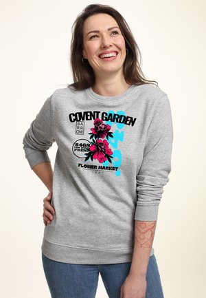Lächelnde Frau mit langen braunen Haaren, die einen grauen Sweatshirt mit rosa Blumen und schwarzem sowie blauem "Covent Garden flower market" Schriftzug trägt.