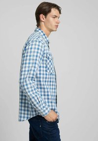 Camicia a quadri blu e bianca con maniche lunghe, taschino sul petto e vestibilità comoda, abbinata a jeans scuri.