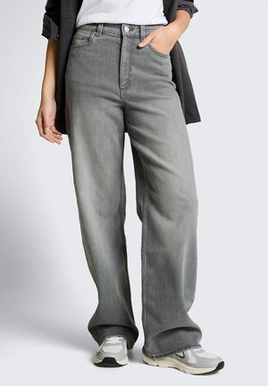 Persoon die losse grijze spijkerbroek draagt met witte sneakers, hand in de zak, en een zwart jasje over een wit topje tegen een effen achtergrond.