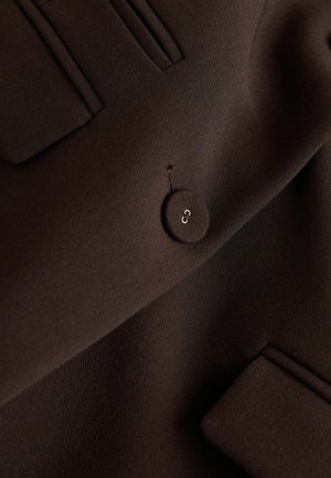 Close-up of dark brown fabric with a round, matching button sewn with two white threads and two welt pockets.