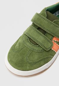 Grønn semsket joggesko med to borrelåsstropper, oransje detaljer, meshfôr, hvit gummisåle og teksturert detaljering på overdelen.