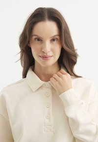 Kremowy sweter polo z kołnierzykiem zapinanym na guziki, ściągaczami na rękawach i miękką teksturą. Ma trójkowy detal w przeszyciach.