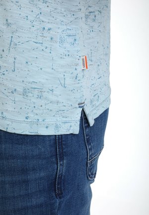 Camisa de manga corta azul claro con un patrón texturizado, presenta una abertura lateral. Etiqueta con el logotipo en el lado izquierdo cerca del dobladillo. Llevada con pantalones de mezclilla.