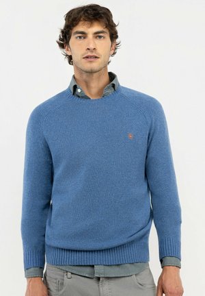 Hombre que lleva un suéter azul claro sobre una camisa gris abotonada y pantalones grises, de pie frente a un fondo blanco liso.