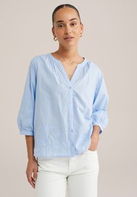 Light blue blouse with a V-neck, featuring buttons, puffed sleeves, and intricate floral embroidery along the hem and cuffs. Smooth cotton fabric.