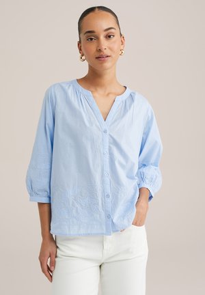 Lyseblå bluse med V-hals, med knapper, puffede ærmer og detaljeret blomsterbroderi langs kanten og manchetterne. Glat bomuldsstof.