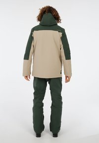 Béžová a zelená lyžiarska bunda s kapucňou, s farebným blokovým dizajnom, nastaviteľnými manžetami a vodeodolnou tkaninou. V páre so zelenými lyžiarskymi nohavicami.