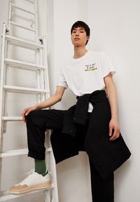Witte t-shirt met zwarte en gele tekst, zwarte broek, groene sokken en witte sneakers met tan kleurige zolen. De pose omvat een witte ladder.