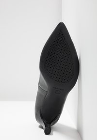 Chaussure noire à talon pointu avec semelle texturée et respirante reposant contre l'angle blanc du mur et du sol.