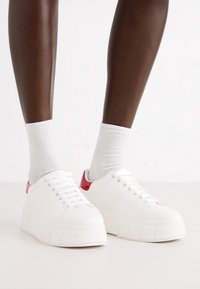 Weiße Platform-Sneaker mit glatter Textur, ausgestattet mit einem roten Fersenakzent und sechs Ösen für Schnürsenkel, kombiniert mit weißen gerippten Socken.