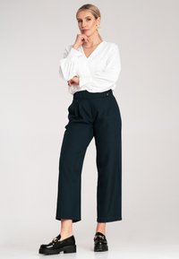 Biała bluzka z długim rękawem i dekoltem w serek, zestawiona z ciemnozielonymi spodniami o wysokim stanie i szerokimi nogawkami oraz czarnymi butami na platformie z złotymi akcentami.