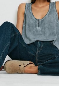 Grå ærmeløs top med rund halsudskæring og knaplukning, kombineret med mørkevidde cowboybukser og beige ruskindssandaler med rem.