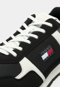 Black sneaker with white laces and accents, featuring a rubber sole, fabric upper, and a logo patch displaying red, white, and blue stripes.