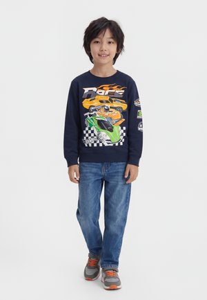 Garçon aux cheveux foncés portant un sweat-shirt bleu marine avec des motifs de voitures de course, un jean bleu et des baskets grises avec des lacets orange, debout sur fond blanc.