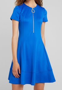 Ljusa blå klänning i en mjuk väv. Har korta ärmar, rund halsringning och en framdragkedja med ett cirkulärt draghandtag. Har en utsvängd kjol.