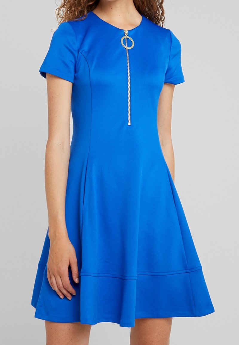 Ljusa blå klänning i en mjuk väv. Har korta ärmar, rund halsringning och en framdragkedja med ett cirkulärt draghandtag. Har en utsvängd kjol.