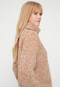 Udoben velik pleten pulover v oranžni in kremni mešanici, ki ima visok ovratnik in teksturiran vzorec po tkanini.