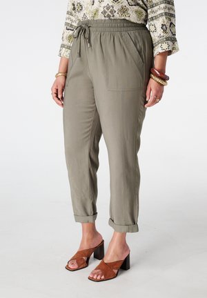 Vrouw draagt beige broek met trekkoord en opgerolde pijpen, een blouse met lange mouwen en patroon, bruine sandalen met hak, en dikke armbanden.