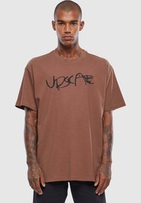 Brun oversized t-shirt lavet af bomuld. Har et fremtrædende sort grafisk print over brystet og korte ærmer.