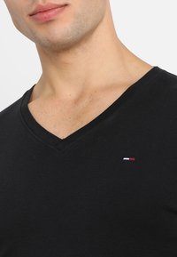 Schwarzes Baumwoll-V-Ausschnitt-T-Shirt mit einer figurbetonten Passform. Hat ein kleines gesticktes Logo in Rot, Weiß und Blau in der Nähe des Ausschnitts.