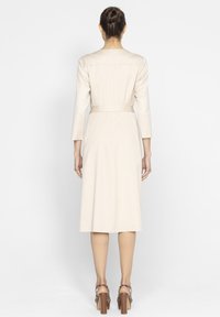 Beżowa sukienka z paskiem, z długim rękawem i prostym krojem, o gładkiej fakturze i długości do kolan, w zestawieniu z szpilkami.