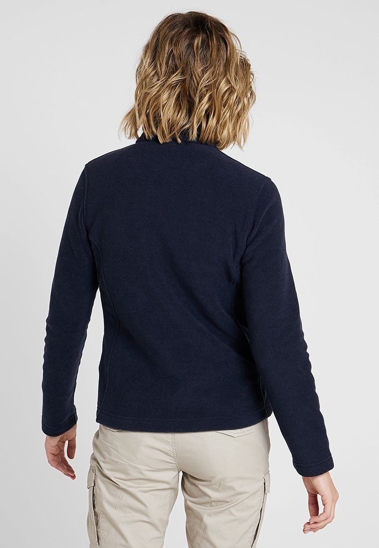 Personne aux cheveux blonds ondulés mi-longs portant un haut à manches longues bleu marine et un pantalon cargo beige, de dos sur un fond uni.