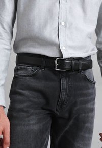 Cinturón de cuero negro con hebilla de metal plateado, usado sobre unos jeans de mezclilla gris y una camisa de botones gris claro.
