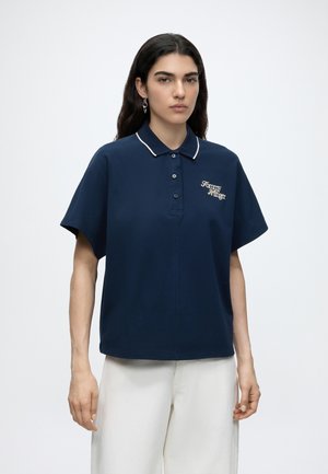Personne aux longs cheveux foncés portant un polo bleu marine avec un passepoil blanc et un logo brodé blanc, associé à un pantalon blanc ample.