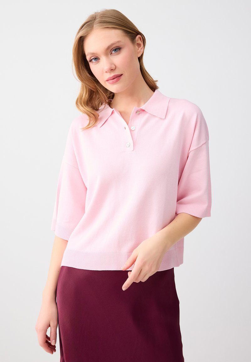 Femme aux cheveux châtain clair portant une chemise à manches courtes rose clair avec col et une jupe bordeaux foncé sur un fond uni.
