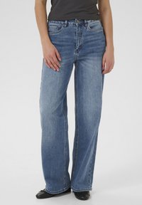 Persona che indossa jeans a vita alta, larghi, di colore blu con lavaggio sbiadito, ballerine nere e una maglietta nera, in piedi contro uno sfondo neutro.
