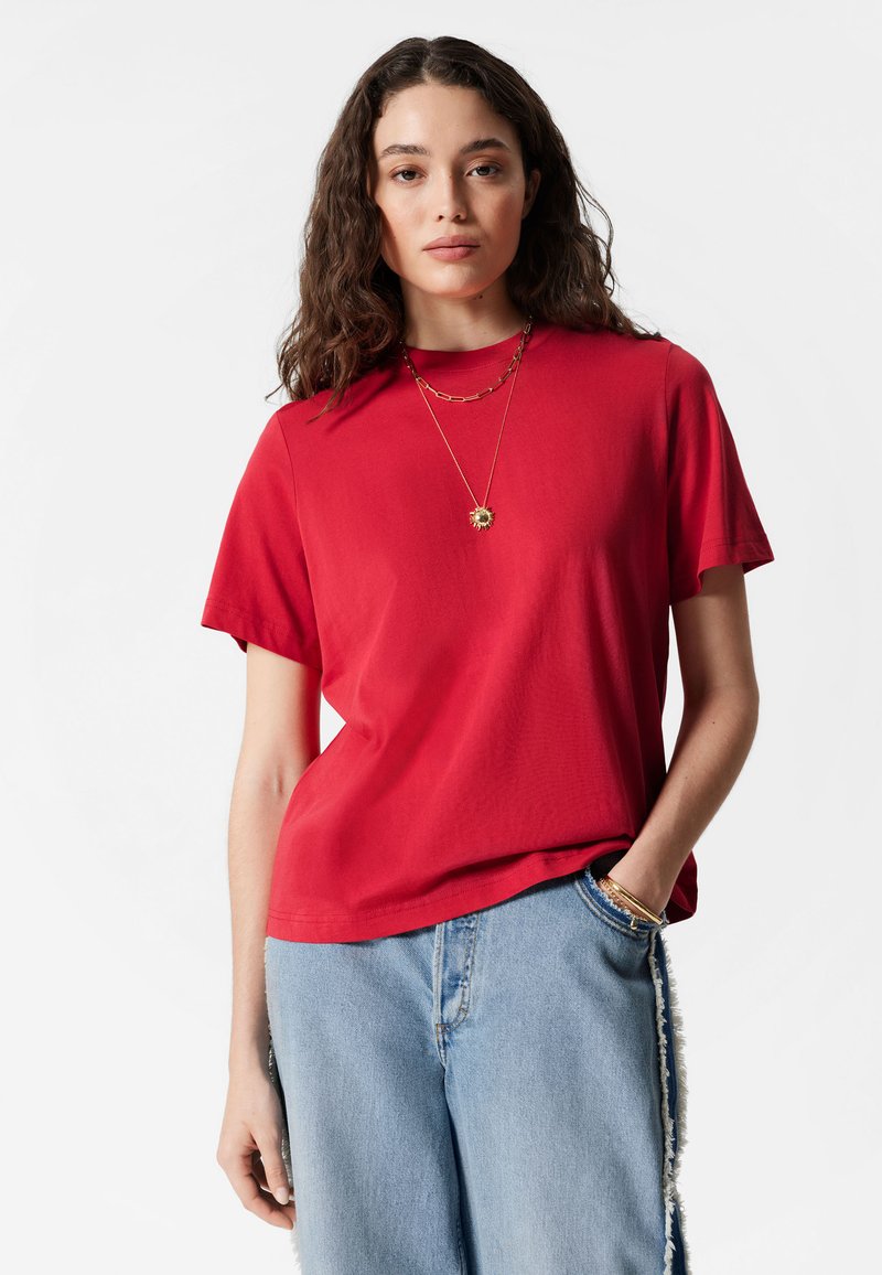 Rotes Baumwoll-T-Shirt mit Rundhalsausschnitt und kurzen Ärmeln, kombiniert mit hellblauen Jeans aus Denim, die Fransenakzente entlang der Nähte haben.