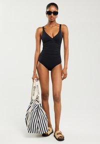 Melna viengabala peldkostīma ar pārklāšanas dizainu un plānām siksniņām, apvienota ar svītrainu somu un bēšu sandālēm. Gluda tekstūra, šaura piegriezte.