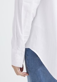 White long-sleeve shirt with vertical pinstripes, button cuff detail, and a rounded hem, paired with blue denim jeans.