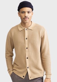 Beige strikket cardigan med et teksturert mønster, som har en kontrastfarget kremfarget krage, seks sorte knapper og ribbestrikkede mansjetter. Brukt over rutete bukser.