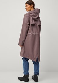 Trinchera de color malva, resistente al agua, con capucha ajustable y ventilación en la parte posterior. Combinado con botas oscuras y jeans de mezclilla azul.