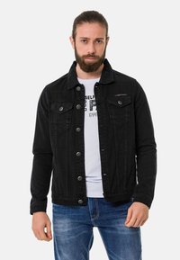 Veste en denim noir avec deux poches poitrine, fermeture à boutons et coupe droite. Portée sur un t-shirt graphique blanc et un jean bleu.
