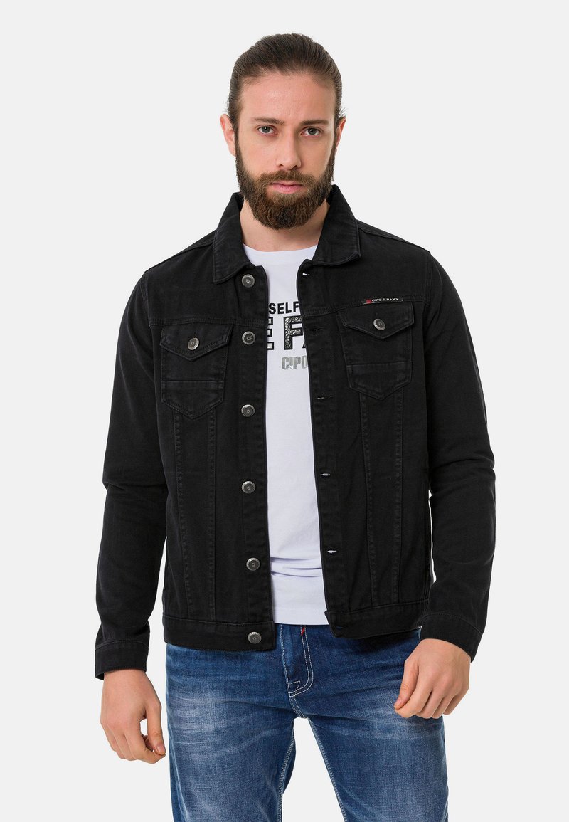 Veste en denim noir avec deux poches poitrine, fermeture à boutons et coupe droite. Portée sur un t-shirt graphique blanc et un jean bleu.