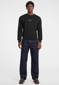 Felpa nera con collo rotondo e logo, abbinata a jeans dritti blu scuro. L'outfit presenta un taglio casual e rilassato.