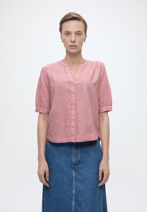 Femme portant un chemisier à manches courtes rose clair à boutons, associé à une jupe en denim bleu taille haute, debout devant un fond uni.