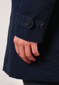 Dłoń spoczywająca obok granatowego rękawa płaszcza z dwoma czarnymi guzikami na regulowanym pasku mankietu.