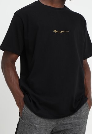 Man wearing a black short-sleeve t-shirt with yellow cursive text and gray plaid pants with hands in pockets.