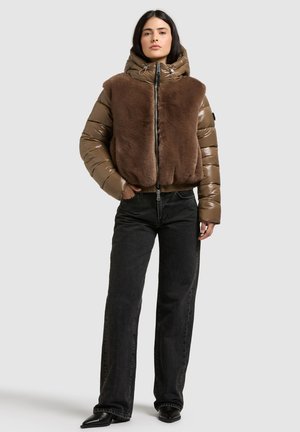 Un mannequin porte une veste marron à capuche avec des empiècements doux sur le devant, un jean noir et des bottes pointues sur un fond clair uni.