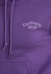 Violetti huppari, jossa on kiristysnyörit; siinä on kenguru-tasku ja valkoinen "Carhartt WIP" -logo rinnassa rohkealla tekstillä. Sileä pinta.