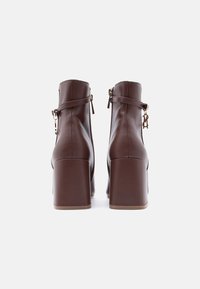 Ζευγάρι καφέ δερμάτινα μποτάκια με χοντρό τακούνι, που φαίνονται από πίσω, με φερμουάρ και ρυθμιζόμενα λουράκια στον αστράγαλο.