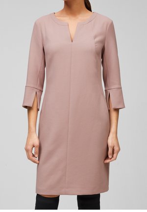 Mujer vistiendo un vestido malva a la altura de la rodilla con mangas tres cuartos y aberturas frontales en los puños, de pie contra un fondo gris liso.