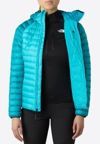 Lätt, turkos puffjacka med huva och dragkedjestängning; quiltad textur; bärs över en svart långärmad topp.