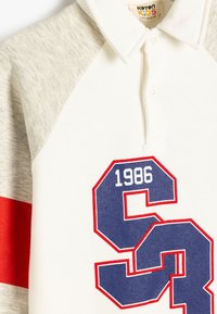 Camisa polo blanca y gris con un acento rojo. Presenta un número "1986" en azul y rojo y el logo "S". Está hecha de una tela suave con un diseño de cuello.