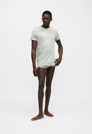 Homme debout pieds nus portant un t-shirt gris clair à manches courtes et un sous-vêtement assorti sur fond blanc uni.